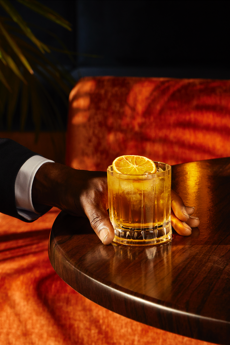 Hand reaching for a non-alcoholic Old Fashioned recipe in a glass with an orange slice on a wooden table.