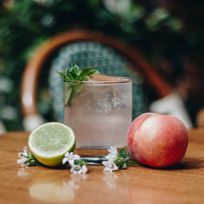 Yasmine cocktail served with fresh mint, peach, lime, and flowers on a wooden table.